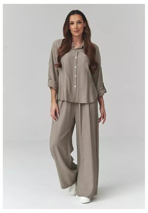 Kobieta stojąca, ubrana w luźną taupe koszulę na guziki z podwiniętymi rękawami oraz dopasowane szerokie spodnie, białe buty, na jednolitym tle.