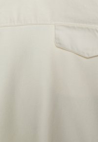 Tessuto di cotone bianco con superficie strutturata, caratterizzato da una singola tasca sul petto e dettagli di cuciture visibili. Presenti leggere variazioni di colore.