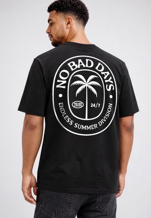 Mann trägt schwarzes T-Shirt mit weißem kreisförmigen Palmen-Grafik und Text „NO BAD DAYS, NEVER LESS, 24/7, ENDLESS SUMMER DIVISION“ auf dem Rücken.