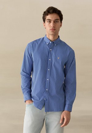 Noormees tumedate juustega, kellel on seljas sinise ja valge vertikaalsete triipudega nööbitav triiksärk ning helesinised teksad, seisab ühe käega taskus.