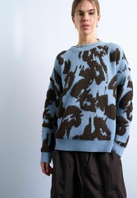 Jeune femme portant un pull bleu avec de grands motifs abstraits texturés marron foncé et un pantalon ample foncé, sur un fond clair uni.