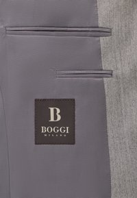 Šedá látka s rybovou kostrou má dve rovnobežné vrecká na ľavej strane. Na látke je zošité čierne označenie s nápisom "BOGGI MILANO" v bielej farbe.