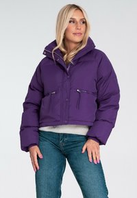 Krojona fioletowa kurtka puchowa wykonana z błyszczącego materiału, z wysokim kołnierzem, dwiema zapinanymi na zamek kieszeniami z przodu i zapięciem na guziki.
