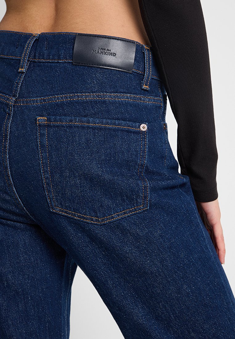 Gros plan sur une personne portant un jean en denim bleu foncé avec un haut à manches longues noir, la main reposant près de la cuisse, poche arrière visible.