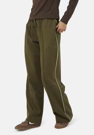 Personne portant un pantalon large vert olive avec des bandes blanches sur les côtés et des baskets marron, debout avec une main dans la poche.