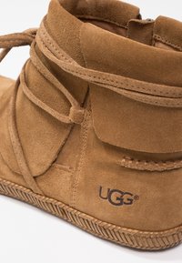 Bottine en daim marron avec lacets enroulés, coutures visibles, semelle texturée et logo UGG sur la zone du talon.