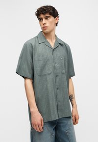 Νέος άνδρας που φοράει γκρι κοντομάνικο πουκάμισο με δύο τσέπες στο στήθος, σε συνδυασμό με φαρδιά μπλε τζιν, κοιτάζοντας προς τα πλάγια.