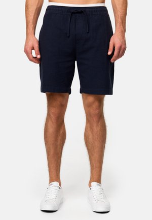 Shorts en lin bleu marine avec taille élastique et cordon de serrage, dotés d'une coupe décontractée et d'ourlets roulés, portés avec des baskets blanches.
