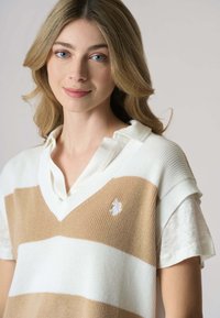 Maglione a collo a V a righe beige e bianche, con maniche corte e finitura testurizzata, dotato di un piccolo emblema sul petto.