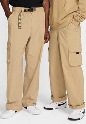 Beige cargo bukser med flere lommer, elastisk talje og mønstret bælte. Bæret med hvide sneakers med sorte accenter.