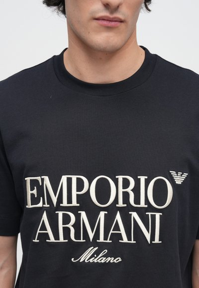 Μαύρο βαμβακερό T-shirt με μεγάλα λευκά γράμματα "EMPORIO ARMANI" και "Milano". Περιλαμβάνει έμβλημα λογοτύπου στο πάνω δεξί μέρος.