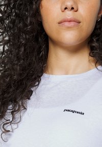 White cotton t-shirt with round neckline, featuring a black "patagonia" logo embroidered on the left chest. Curly hair partially visible.