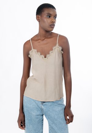 Femme portant un caraco beige orné de dentelle et un jean bleu clair, debout devant un fond blanc uni, regardant sur le côté.