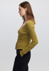 Cardigan a coste verde senape con scollatura a V profonda, maniche lunghe e chiusura con bottoni frontali. Tessuto testurizzato con una silhouette aderente.