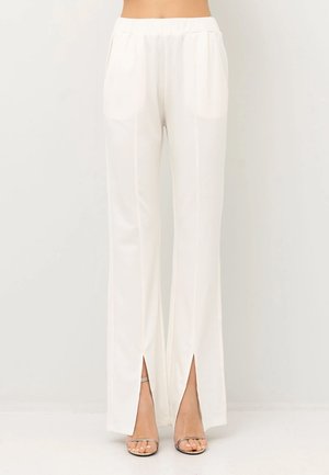 Pantaloni bianchi a gamba larga con una texture liscia, caratterizzati da spacchi laterali, una cintura elastica e due tasche anteriori. Abbinati a sandali trasparenti con cinturini.