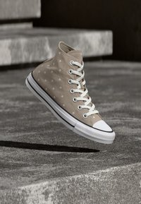 Μπεζ ψηλό αθλητικό παπούτσι με λευκά αστέρια και κορδόνια που αιωρείται πάνω από μια τσιμεντένια επιφάνεια με σκιά από κάτω.