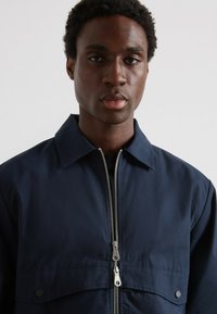 Giacca blu navy con colletto piatto, chiusura a zip e tasche anteriori. Tessuto morbido con una leggera lucentezza e dettagli di cucitura accentuati.