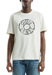 Jonge man in een off-white T-shirt met zwarte ronde tekstgrafiek "QUIET SENSE", gecombineerd met blauwe jeans, staand tegen een witte achtergrond.