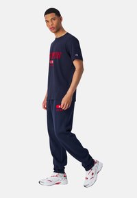 Navy blauwe katoenen T-shirt met rode "CHAMPION" tekst, gecombineerd met bijpassende navy joggers. Beide hebben logo-accenten; model draagt witte sneakers.