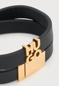 Μαύρο δερμάτινο βραχιόλι με διπλό λουράκι που διαθέτει χρυσούς χαρακτήρες "HUGO" και ορθογώνιο χρυσό διακοσμητικό. Λείο υλικό με αντίθετα στοιχεία.