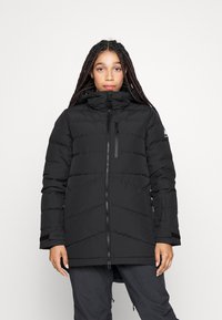 Zwarte pufferjas met een hoge kraag, voorrits en diagonale quiltontwerp. Beschikt over ritssluitingen aan de zijzakken en klittenbandaccenten op de mouwen.
