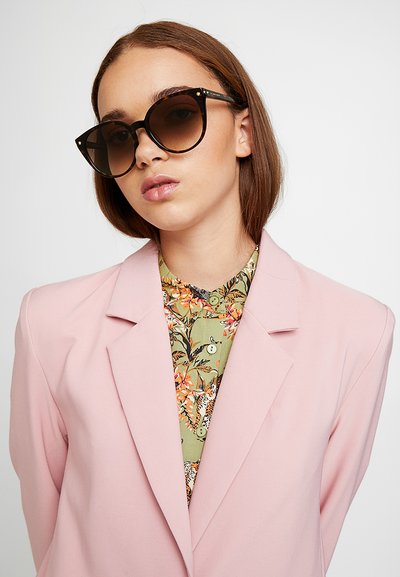Sorte cat-eye solbriller med mørke linser, kombinert med en bluse med blomstermønster og en lys rosa blazer med en strukturert design.