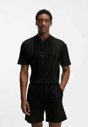 Mężczyzna ubrany w czarną koszulkę z krótkim rękawem i pasujące szorty z subtelnymi pionowymi paskami, stoi z jedną ręką w kieszeni na białym tle.