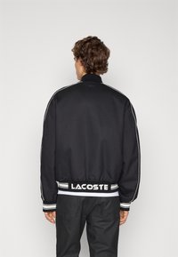 Svart bomberjacka med vita axelränder och ribbade manschetter, med en texturerad överdel och en logotyp på baksidan och midjebandet.