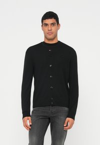 Vīrietis valkā melnu pogātu kardiganu pār melnu T-kreklu un pelēnas džinsas, stāvot pret vienkāršu gaišu fonu.