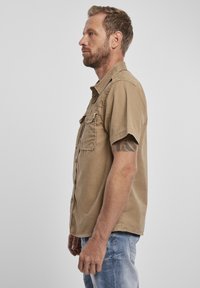 Camicia khaki a maniche corte, con bottoni e due tasche frontali, colletto aperto, realizzata in tessuto testurizzato. Indossata con jeans azzurri chiari.