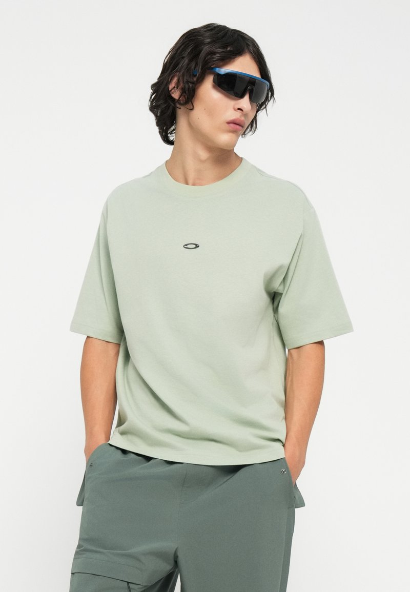 T-shirt vert clair à manches courtes avec un col rond et un petit logo noir sur la poitrine, associé à un short vert foncé et des lunettes de soleil noires.