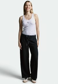 Weißes Tanktop mit einem Logo, kombiniert mit schwarzen weit geschnittenen Hosen mit Taschen und einem Kordelzug. Das Modell trägt helle Sneakers.