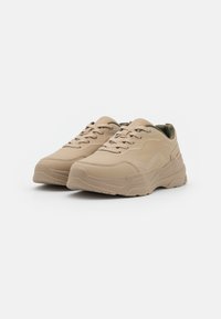 Beige sneakers with a smooth leather upper, lace closure, padded collar, and textured rubber sole. Features contrasting stitching and rounded toe.