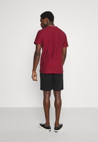 Homme de dos portant une chemise bordeaux à manches courtes, un short noir et des baskets noires et blanches sur un fond uni.