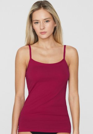 Blondynka o jasnej karnacji, ubrana w dopasowany, bezrękawnik w ciemnoróżowym kolorze, stojąca na tle jednolitego, jasnego tła.