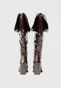 Par de botas de couro castanho-escuras altas, com as partes superiores dobradas e saltos quadrados, mostradas de costas sobre um fundo branco liso.