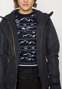 Schwarze Außenjacke mit Reißverschluss und verstellbarer Kapuze, über einem marineblauen Sweater mit einem Bergmuster in Pastellfarben getragen. Matte Textur.