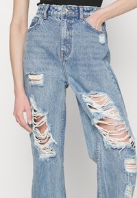 Lys blå denimjeans med høy midje, med flere slitte områder og frynsete kanter. Fem-lomme design, avslappet passform.