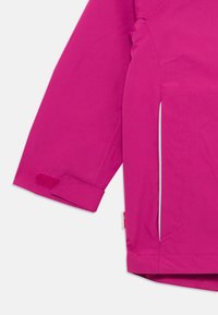 Manga de jaqueta magenta com faixa de velcro ajustável e bolso lateral, mostrando detalhes de costura e uma pequena etiqueta de tecido.