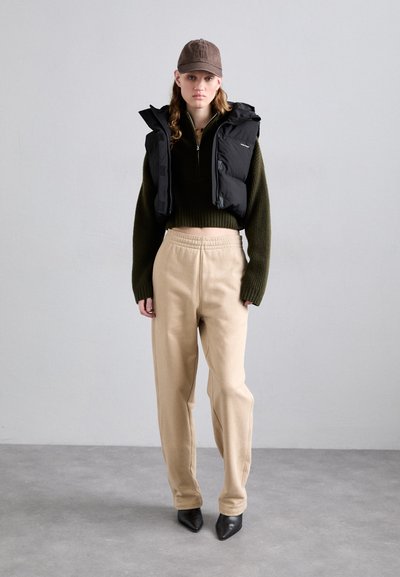 Svart puffer vest over en mørkegrønn cropped genser, beige bukser med elastisk midje og svarte spisse hæler. Brun caps fullfører looken.