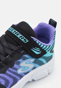Baskets Skechers noires et bleues avec un dessus texturé, des accents violets, une sangle à crochets et une semelle blanche avec un motif.
