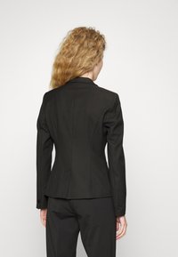 Blazer noir ajusté avec un design sur mesure, manches longues, revers crantés et texture lisse. L'arrière comprend des pinces pour la mise en forme.