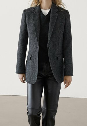 Person wearing a dark grey tailored blazer, black v-neck sweater, white undershirt, and black leather pants, standing against a plain background.