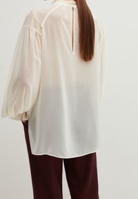 Cream-colored blouse with long puff sleeves, textured braiding along the shoulders, and a back keyhole opening, paired with maroon pants.