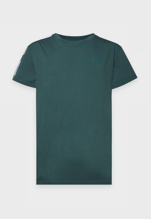 Un homme pose pieds nus, vêtu d’un tee-shirt turquoise à manches courtes et d’un pantalon de jogging noir, devant un fond blanc uni de studio.