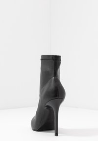 Czarne botki na wysokim obcasie z wąskim noskiem i gładkim wykończeniem, wyeksponowane w pozycji stojącej na białej powierzchni w białym tle w rogu.