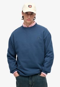Sudadera azul con puños y cuello acanalados, logo en el pecho; llevada sobre una camisa de cuello ligera en color rosa pálido. Gorra beige con detalle de parche.