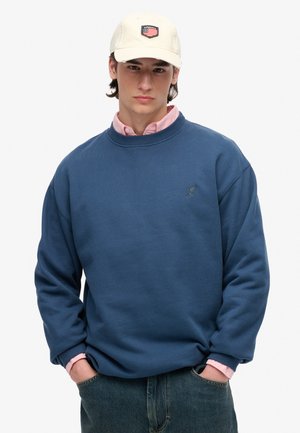 Niebieska bluza z ribbowymi mankietami i dekoltem, z logo na piersi; noszona na jasno różowej koszuli z kołnierzykiem. Beżowa czapka z naszywką.