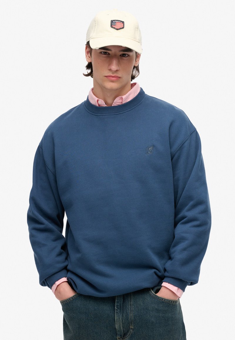 Sudadera azul con puños y cuello acanalados, logo en el pecho; llevada sobre una camisa de cuello ligera en color rosa pálido. Gorra beige con detalle de parche.