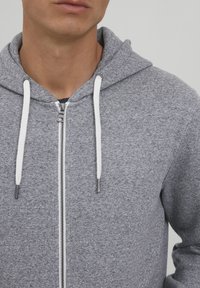 Svijetlosiva hoodie na zatvarač s bijelim vezicama. Ima mekanu teksturu i uski kroj, dizajnirana s visokim ovratnikom i metalnim zatvaračem.